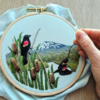 Embroidery hoop with a scenic design of birds, plants, and mountains in the background. 