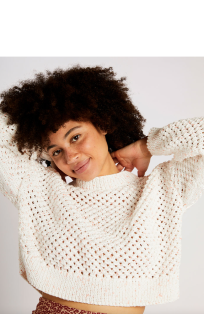 Model with curly brown hair wearing a white eyelet knit sweater on a light gray background.