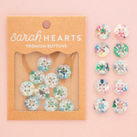 Sarah Hearts Premium Buttons in kraft paper packaging, shown next to ten loose round buttons with iridescent pastel patterns on a soft pink background.