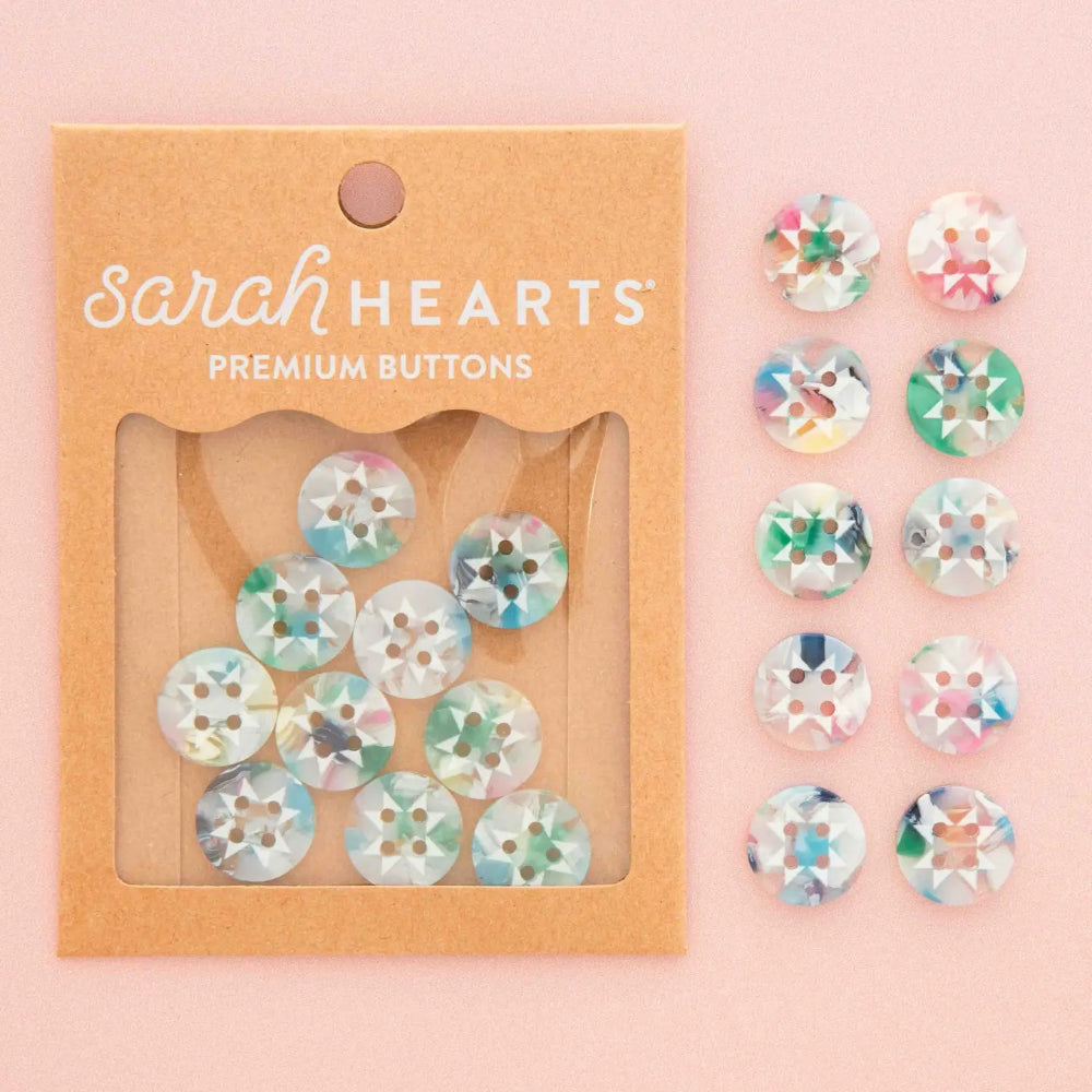 Sarah Hearts Premium Buttons in kraft paper packaging, shown next to ten loose round buttons with iridescent pastel patterns on a soft pink background.