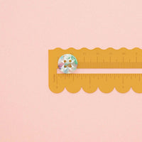 A small round button with a marbled background in pastel blue, green, and pink rests on a yellow scalloped ruler against a soft pink background.