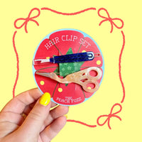 A hand  holds a round card labeled "Hair Clip Set" featuring a blue seam ripper clip and pink stork scissors clip against a yellow background with red bows. 
