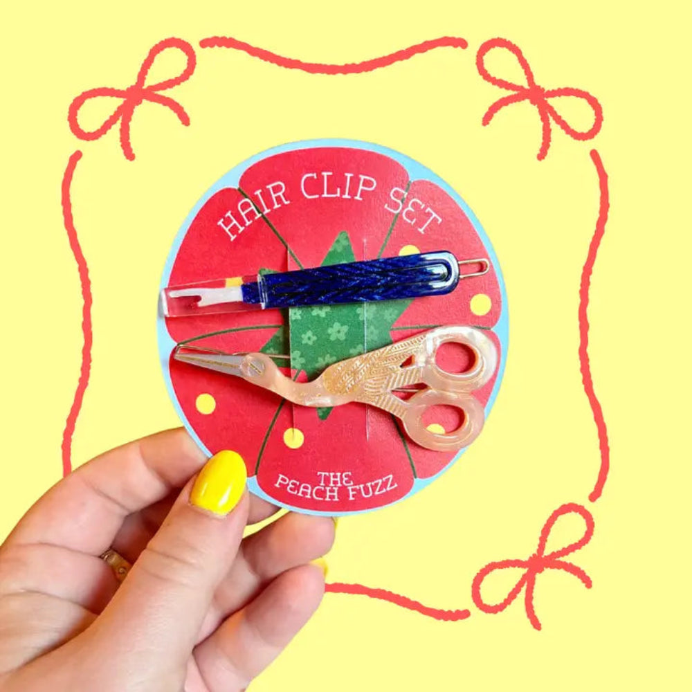 A hand  holds a round card labeled "Hair Clip Set" featuring a blue seam ripper clip and pink stork scissors clip against a yellow background with red bows. 