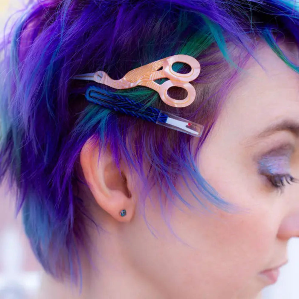 A person with blue and green hair wears a blue seam ripper clip and pink stork scissors clip. 