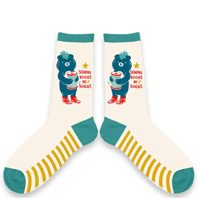 Two socks, each featuring a blue bear with a mohawk and red sneakers holding a big spool of thread with the text 'Sewing rocks my socks' on a sock of blue and white with yellow stripes