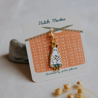 A gold-tone enamel pin shaped like a ghost on a white display card labeled 'Stitch Marker,' photographed on a soft white fabric surface with a small gray stone and dried flowers nearby.