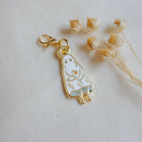 A gold-tone enamel charm shaped like a ghost holding a flower, displayed on a textured off-white fabric next to dried flowers.