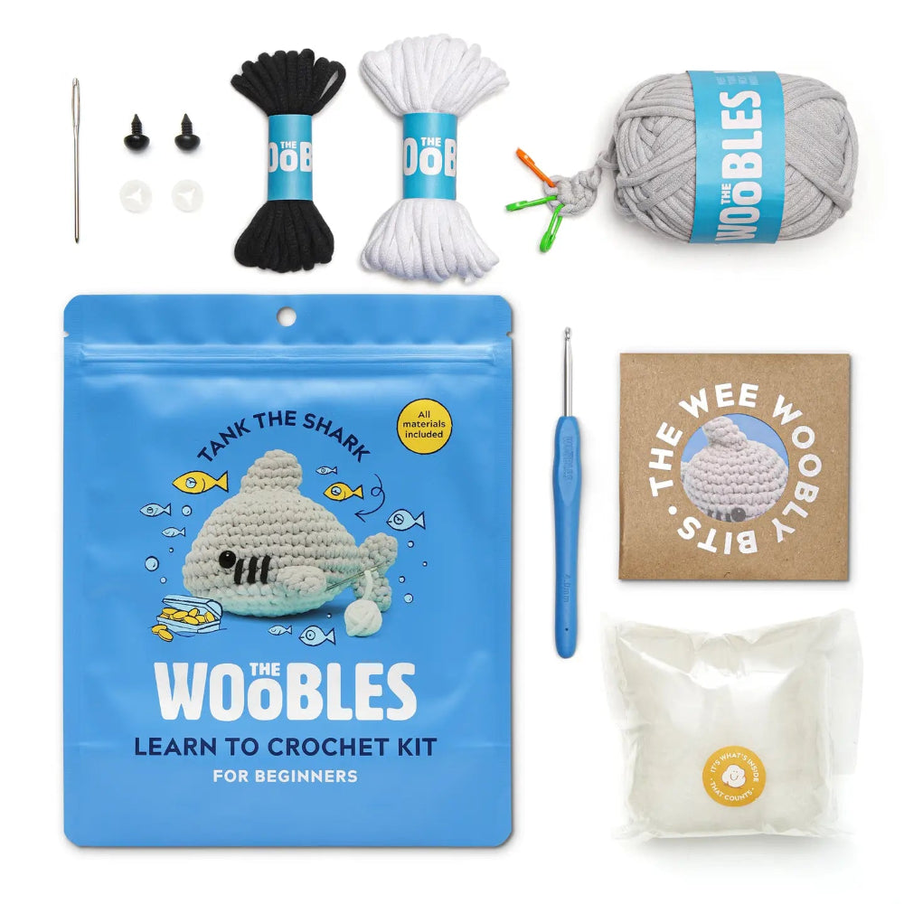 The Woobles Learn to Crochet Kit for Beginners, featuring a blue pouch with a crocheted shark photo, black and white yarn, a gray yarn ball, a blue crochet hook, a small booklet, and other accessories on a white background.