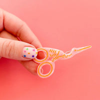 A hand with decorated fingernails holds a small, translucent pink stork scissor-shaped hair clip against a solid pink background.