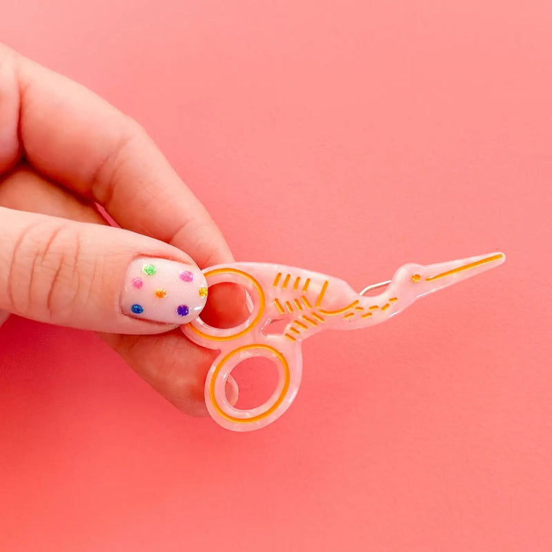 A hand with decorated fingernails holds a small, translucent pink stork scissor-shaped hair clip against a solid pink background.