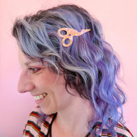A person with lavender and gray wavy hair wearing a translucent pink scissors hair clip, shown in profile against a solid pink background.