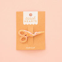 A pink and gold enamel hair clip shaped like a pair of stork scissors, mounted on a brown kraft display card with the brand name 'Sarah Hearts' printed at the top, set against a solid pale pink background.