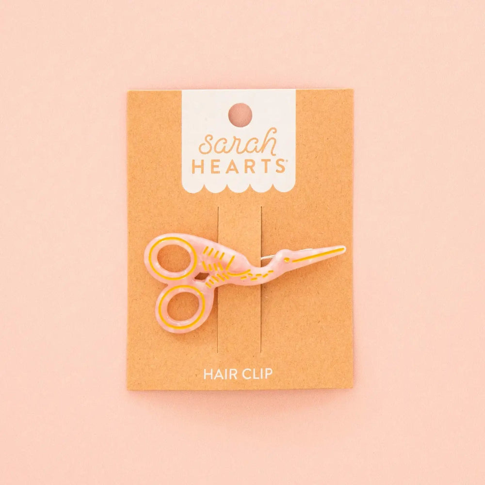 A pink and gold enamel hair clip shaped like a pair of stork scissors, mounted on a brown kraft display card with the brand name 'Sarah Hearts' printed at the top, set against a solid pale pink background.