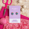 Wooden earrings on a purple card with pink scissors and yarn on a wooden surface