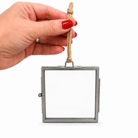 A hand with red nail polish holds a square metal picture frame with a hanging rope loop against a plain white background.