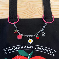 Black tote bag with colorful charm bracelet on a wooden surface, featuring 'Brooklyn Craft Company' branding.