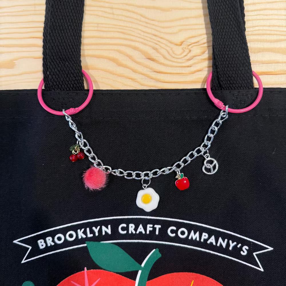 Black tote bag with colorful charm bracelet on a wooden surface, featuring 'Brooklyn Craft Company' branding.