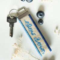 A  cream colored woven fabric keychain with a silver metal keyring and key, with blue needlepointed text reading 'slow down' and blue border stripes. The keychain is styled on a white surface with blueberries and dried flowers.