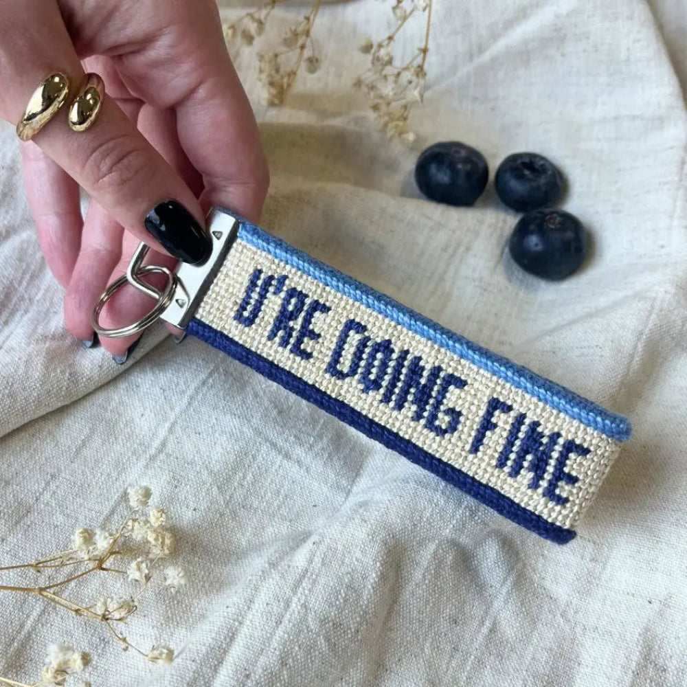 A close-up of a hand holding a needlepoint keychain with the phrase 'U'RE DOING FINE' stitched in navy blue on a cream canvas background, with blue borders. The keychain is displayed on a textured beige linen fabric with dried flowers and blueberries as props.