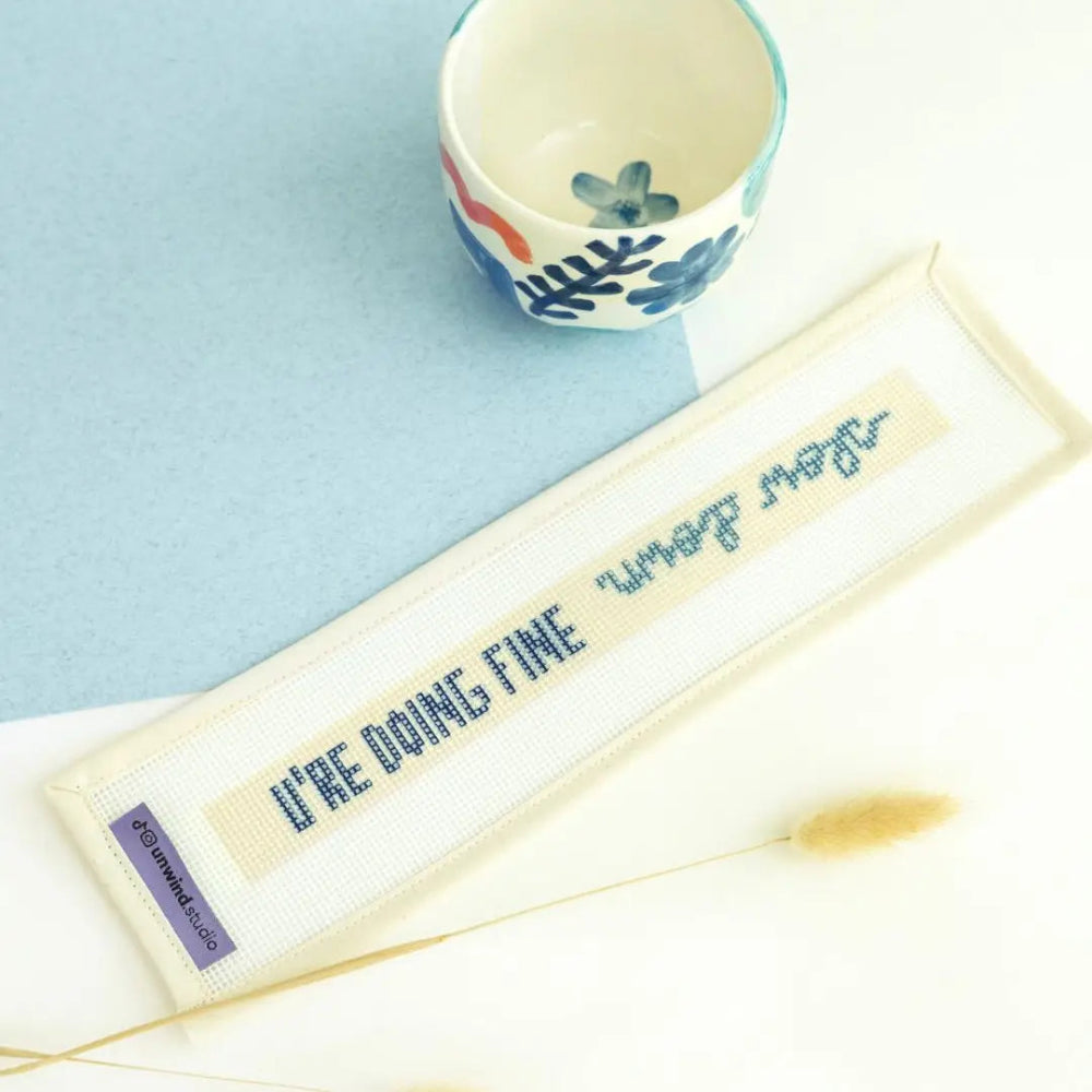 A needlepoint canvas reading 'U're doing fine' and 'Slow Down" in blue on cream fabric, displayed diagonally on a white surface with a light blue background. A small ceramic cup with blue and red floral patterns sits in the upper right, and a dried grass stem lies near the bottom.