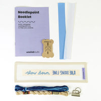 Unwind Studio needlepoint kit laid out on a white background, including a lavender booklet, wooden thread bobbin, blue and white bias tape, cream canvas with blue lettering, blue and tan thread, and a metal binder clip.