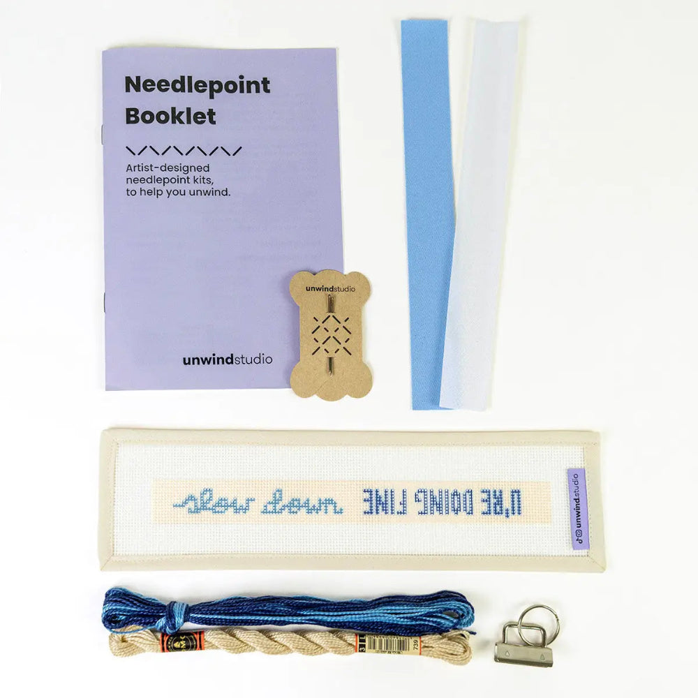 Unwind Studio needlepoint kit laid out on a white background, including a lavender booklet, wooden thread bobbin, blue and white bias tape, cream canvas with blue lettering, blue and tan thread, and a metal binder clip.