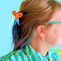 Person with colorful hair and a scissor-shaped hair clip against a blue background