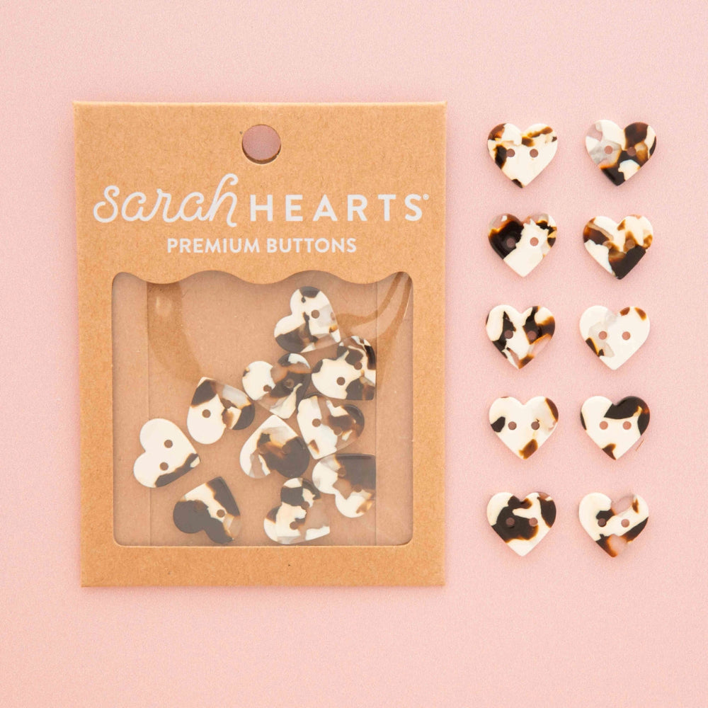Sarah Hearts premium buttons in a tortoise shell print pattern, shown in their kraft paper packaging on the left and ten individual buttons displayed on the right against a solid pink background.