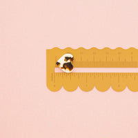 A small tortoiseshell button is shown on a yellow plastic ruler against a solid pale pink background.