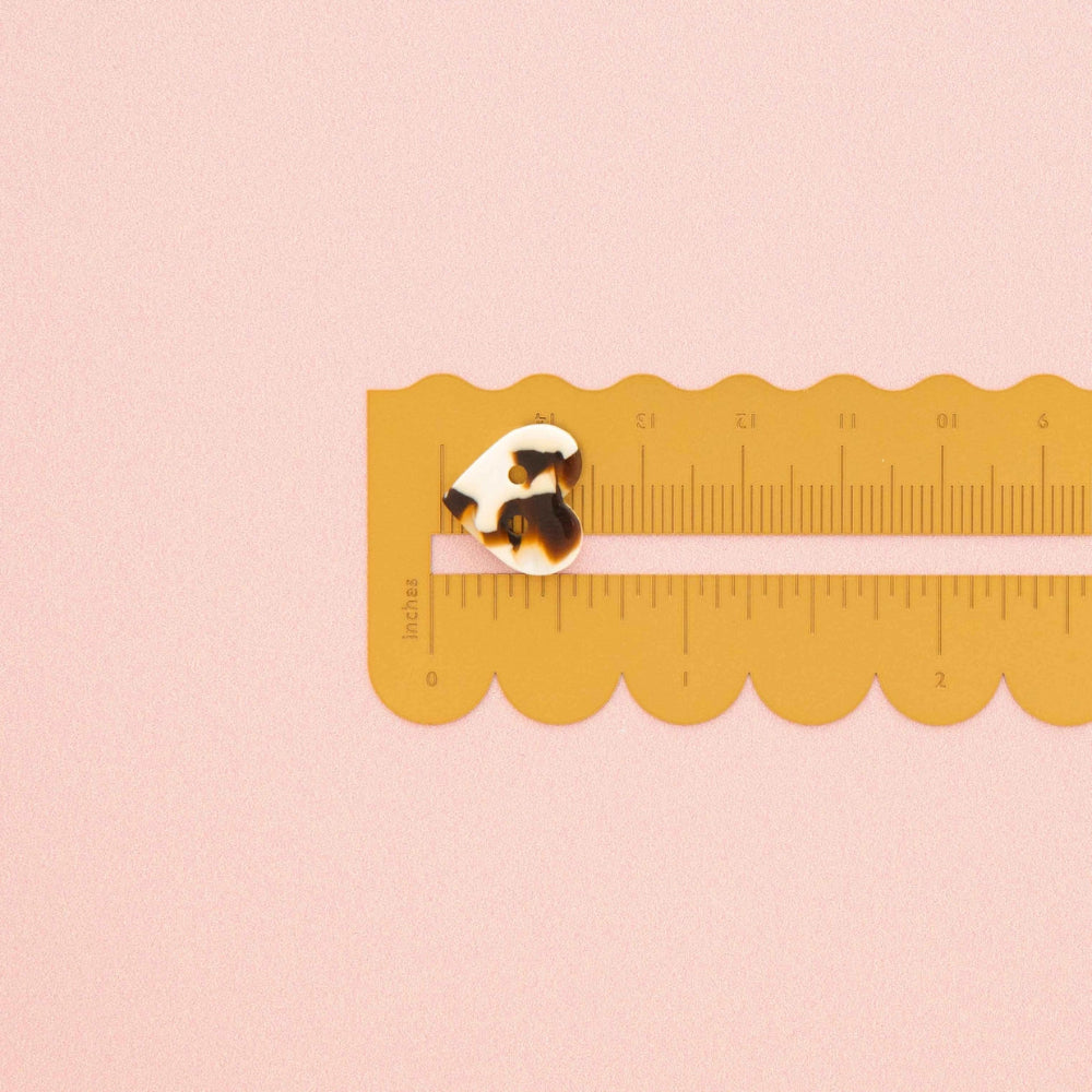 A small tortoiseshell button is shown on a yellow plastic ruler against a solid pale pink background.