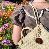 Person holding a woven handbag with a snail keychain attached against a floral background