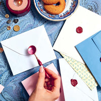 A hand pouring pink wax for a letter seal on a table covered in letters, buttons and colorful paper