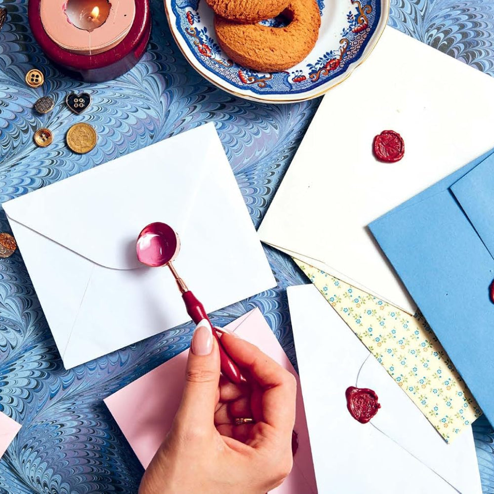 A hand pouring pink wax for a letter seal on a table covered in letters, buttons and colorful paper