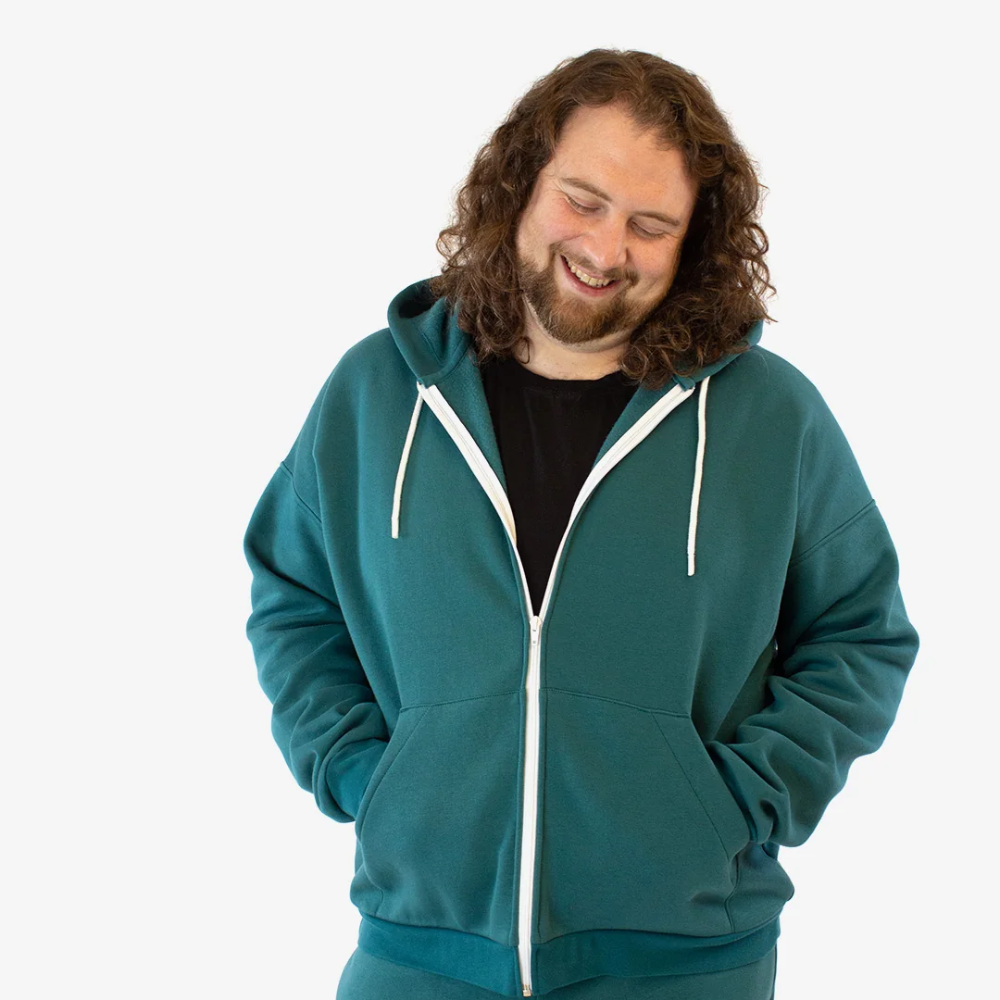 A man in a dark teal zipper hoodie stands smiling with his hands in the pockets