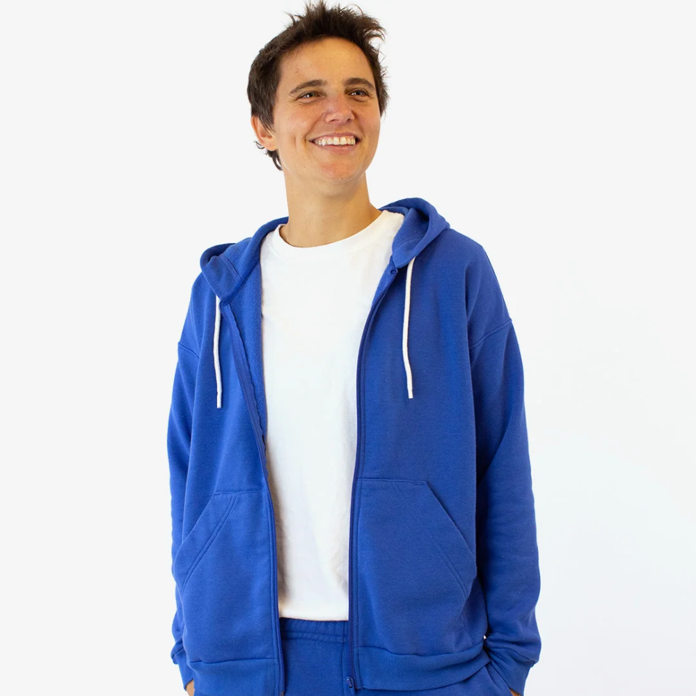 A man in a bright blue zipper hoodie with pockets stands against a white wall
