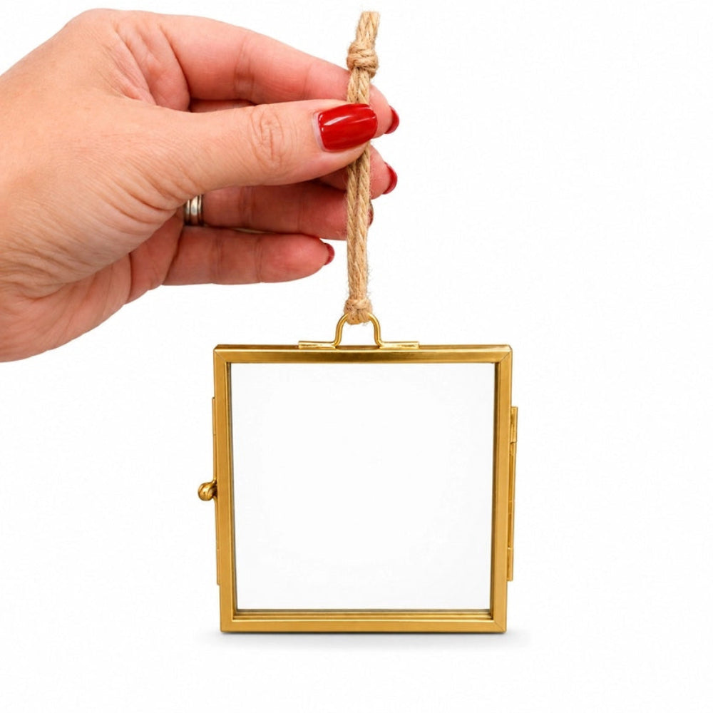 A hand with red nail polish holds a small square gold-framed mirror with a hanging rope against a plain white background.