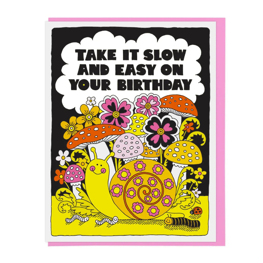A greeting card with a whimsical illustration of a snail, flowers, mushrooms, and caterpillars on a black background, with the text 'TAKE IT SLOW AND EASY ON YOUR BIRTHDAY' inside a white cloud shape. A pink envelope is partially visible behind the card.