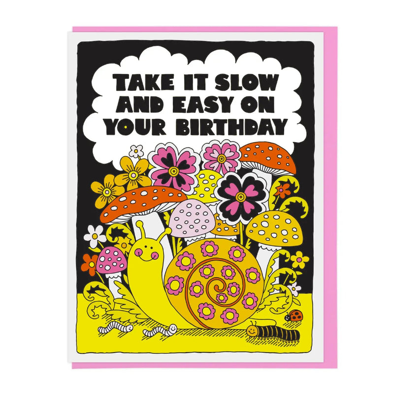 A greeting card with a whimsical illustration of a snail, flowers, mushrooms, and caterpillars on a black background, with the text 'TAKE IT SLOW AND EASY ON YOUR BIRTHDAY' inside a white cloud shape. A pink envelope is partially visible behind the card.