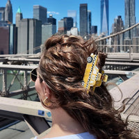 Person with wavy hair wearing a yellow hair clip shaped like a taxi against a city skyline backdrop