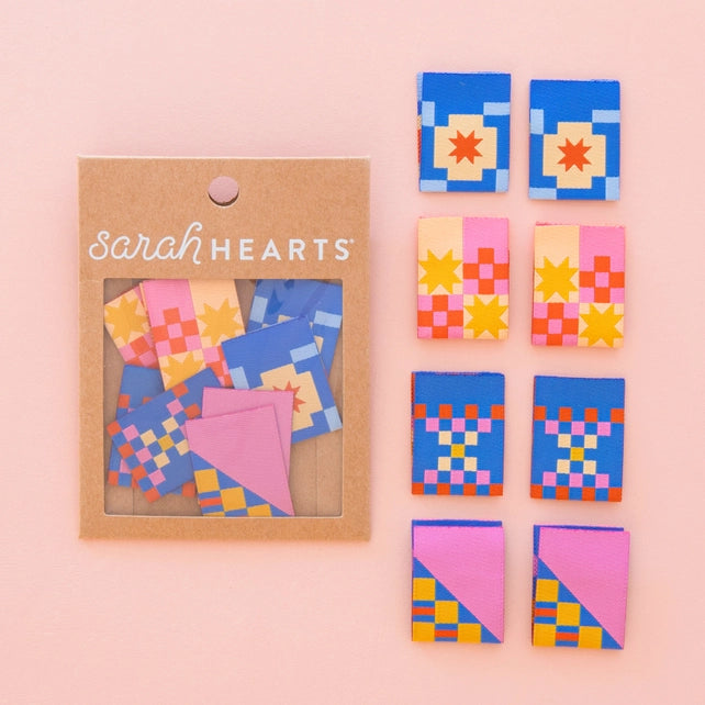 Colorful geometric-patterned quit-style garment labels with 'sarah HEARTS' packaging on a pink background
