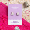 Wooden thimble earrings on a purple card with 'Hawthorn' branding, surrounded by sewing tools on a textured surface.