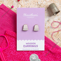 Wooden thimble earrings on a purple card with 'Hawthorn' branding, surrounded by sewing tools on a textured surface.
