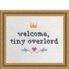 Cross-stitched text 'welcome, tiny overlord' with decorative elements in a gold frame.