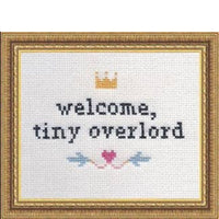 Cross-stitched text 'welcome, tiny overlord' with decorative elements in a gold frame.