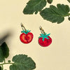 Tomato-shaped earrings with green stems on a beige background with leaf patterns.