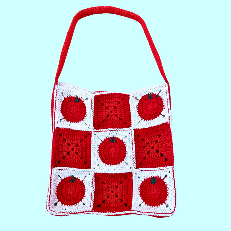 A handmade crocheted shoulder bag featuring a 3x3 grid of red and white granny squares, with three squares decorated as red apples with green stems. The bag has a thick red shoulder strap and is photographed against a solid light blue background.