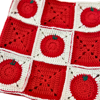 A close-up of a crocheted textile featuring alternating red and white square panels, with several tomato motifs and small green leaf details.