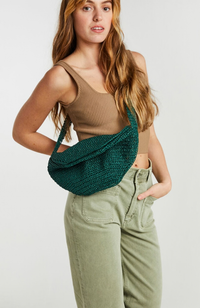 A woman wearing a dark green crocheted crescent shoulder bag over a brown ribbed tank top and sage green pants, photographed against a light gray studio background.