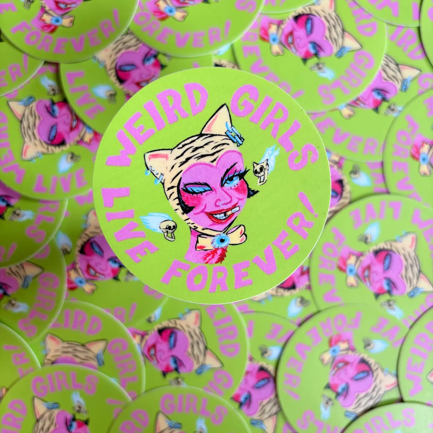 Pile of circular green stickers with the text "Weird girls live forever!" and an illustration of a girl wearing cat ears.