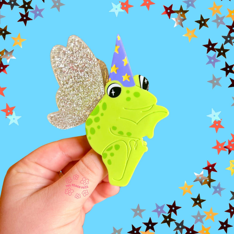 Hand holding a green frog-shaped claw clip with a wizard hat against a blue background with colorful stars.